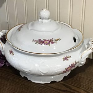 May include: White porcelain soup tureen with lid, featuring gold trim and floral accents in purple and yellow. The tureen has ornate handles and a knob on the lid. A classic piece for serving soups and stews.