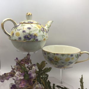 May include: A porcelain tea set featuring a teapot and teacup. The set is decorated with a floral pattern of yellow and purple flowers on a white background. Gold trim accents the edges and handle.