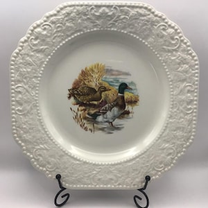 以下が含まれることがあります： 正方形で縁が波状になった白いセラミックプレート。縁には詳細な浮き彫りの花柄模様があり、中央には沼地を背景にした2羽のアヒルの画像があります。アヒルは自然な色で描かれています。