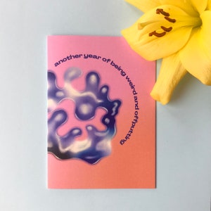 May include: Pink card with a blue and white abstract design and the text "another year of being weird and offputting".