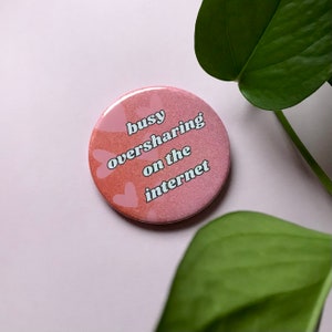 May include: A round, pink button with the text "busy oversharing on the internet" in a white, playful font. The button has a glittery finish and heart-shaped accents.