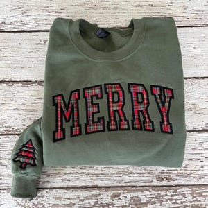 May include: An olive green sweatshirt with the word "MERRY" in a red and black plaid design across the chest. A small plaid Christmas tree is on the left sleeve. The sweatshirt is folded on a white wooden surface.