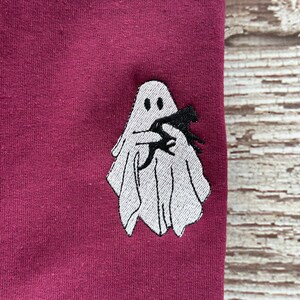 May include: A maroon colored t-shirt with a white embroidered ghost holding a black bat.