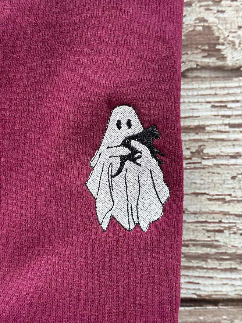May include: A maroon colored t-shirt with a white embroidered ghost holding a black bat.