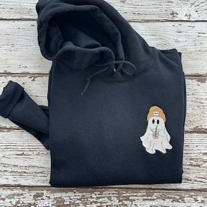 May include: Black hooded sweatshirt with a white ghost wearing a yellow beanie and holding a brown beverage. The ghost has a smiling face.