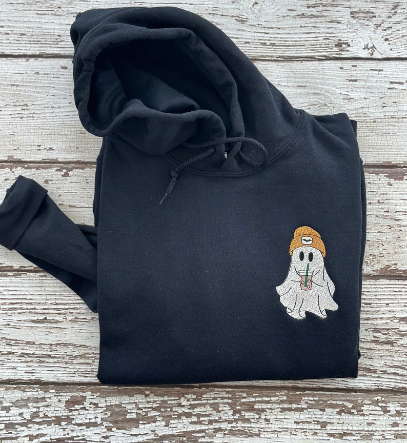 May include: Black hooded sweatshirt with a white ghost wearing a yellow beanie and holding a brown beverage. The ghost has a smiling face.