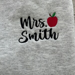 May include: Gray sweatshirt with black embroidered text that reads "Mrs. Smith" and a red apple with a green stem and leaf.