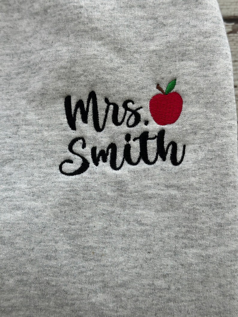 May include: Gray sweatshirt with black embroidered text that reads "Mrs. Smith" and a red apple with a green stem and leaf.