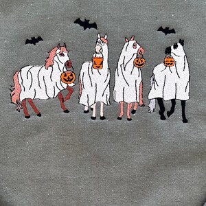 Op de afbeelding: Vier paarden verkleed als geesten voor Halloween, elk met een emmer met een pompoen. De paarden zijn roze en wit met zwarte hoeven en staan ​​op een groene achtergrond met vleermuizen die erboven vliegen.