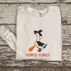 Puede incluir: Sudadera blanca de manga larga con una imagen bordada en negro y naranja de un ganso con un sombrero de bruja y sosteniendo una escoba. El texto "Honkus Ponkus" est&aacute; bordado debajo de la imagen.