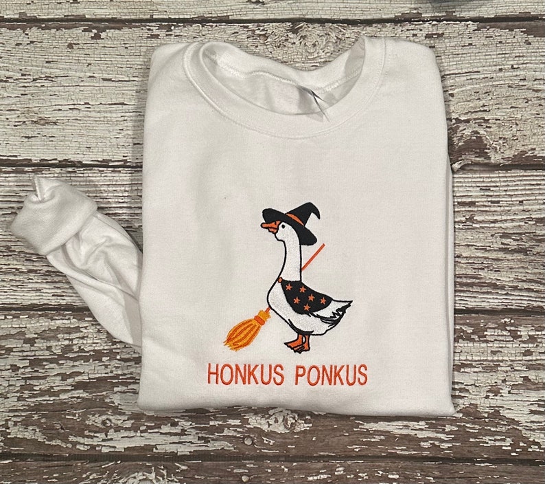 Puede incluir: Sudadera blanca de manga larga con una imagen bordada en negro y naranja de un ganso con un sombrero de bruja y sosteniendo una escoba. El texto "Honkus Ponkus" est&aacute; bordado debajo de la imagen.