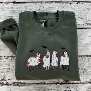 Op de afbeelding: Een groen sweatshirt met een geborduurd ontwerp van vier paarden die verkleed zijn als geesten en emmers met snoep vasthouden. De paarden dragen witte lakens en er vliegen vleermuizen boven hen.
