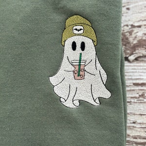 May include: Olive green sweatshirt with an embroidered ghost design. The ghost is wearing a green beanie and holding a coffee drink with a straw. The ghost has two black eyes and a smiling mouth. The design is centered on the sweatshirt.