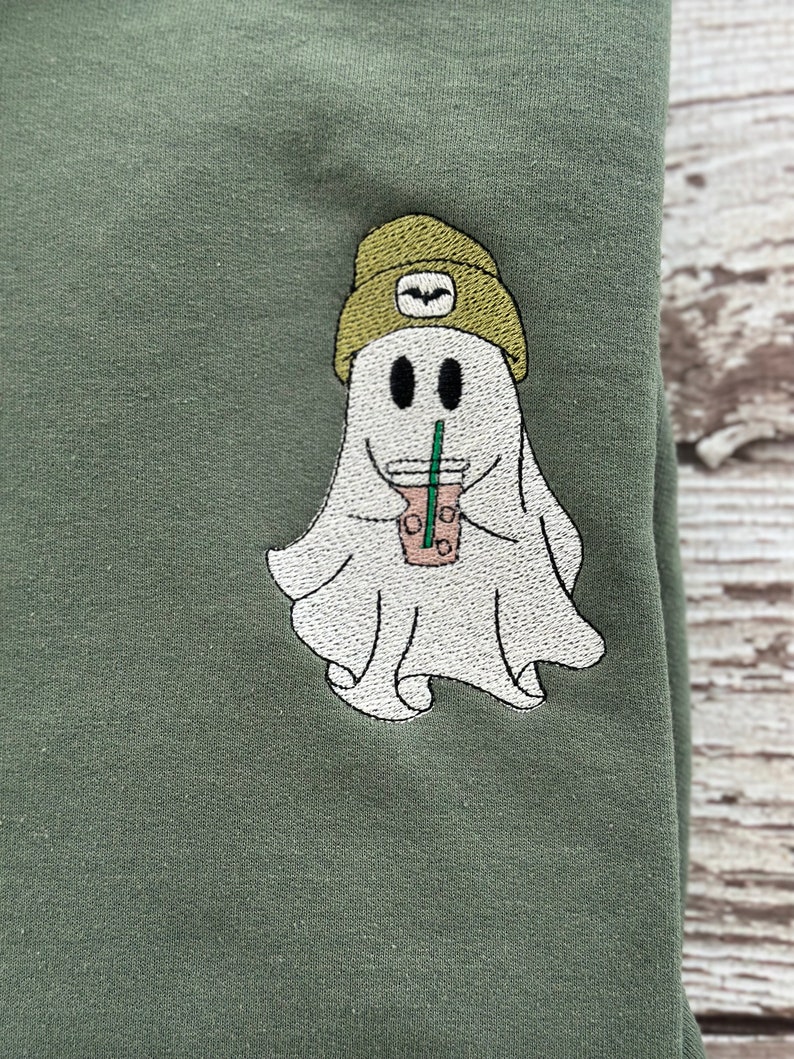 May include: Olive green sweatshirt with an embroidered ghost design. The ghost is wearing a green beanie and holding a coffee drink with a straw. The ghost has two black eyes and a smiling mouth. The design is centered on the sweatshirt.