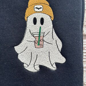 May include: Embroidered design of a ghost wearing a yellow beanie and holding a cup of coffee with a straw. The ghost has two black eyes and a smiling mouth.