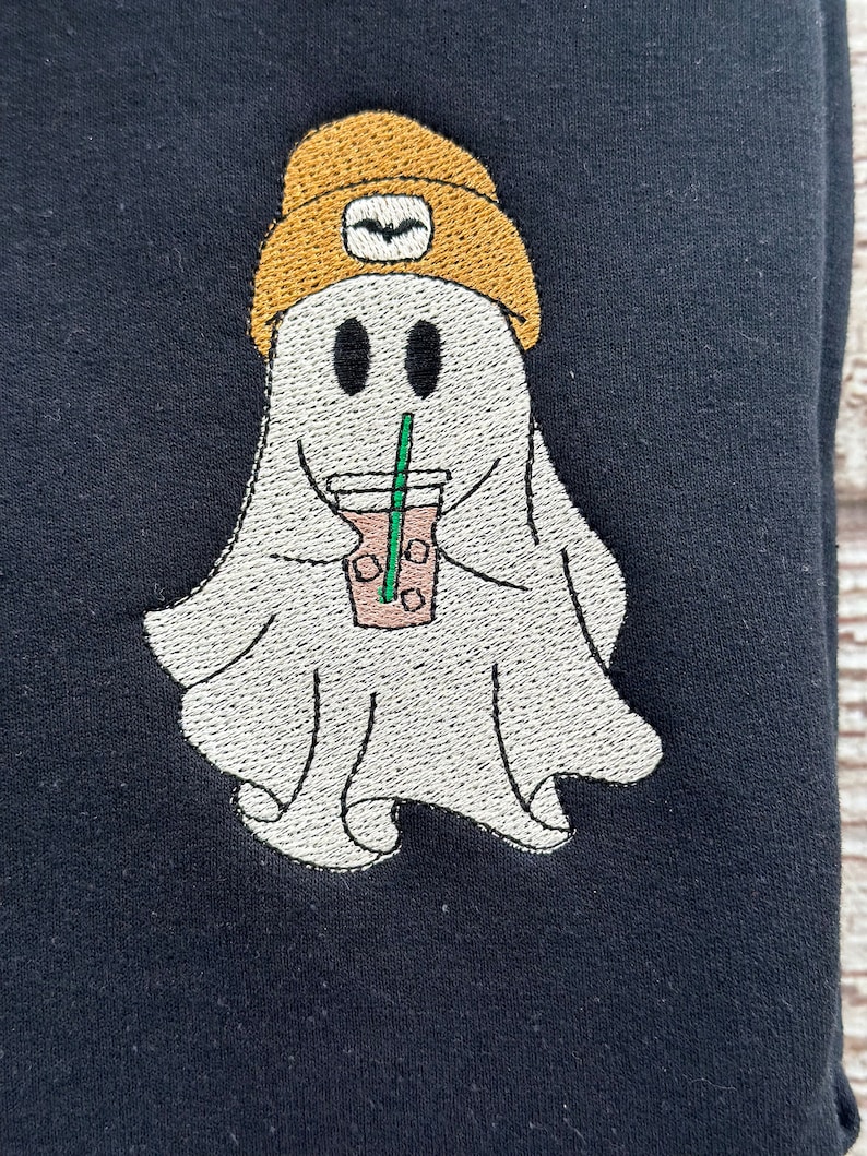 May include: Embroidered design of a ghost wearing a yellow beanie and holding a cup of coffee with a straw. The ghost has two black eyes and a smiling mouth.