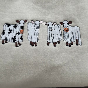 May include: A light beige sweatshirt with four embroidered cows wearing ghost costumes. The cows are holding orange buckets with jack-o'-lantern faces.