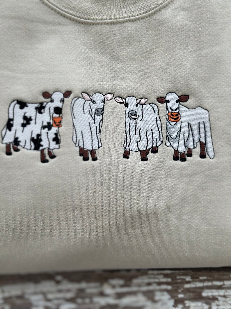 May include: A light beige sweatshirt with four embroidered cows wearing ghost costumes. The cows are holding orange buckets with jack-o'-lantern faces.