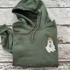 May include: Olive green hoodie with a drawstring hood and a front pocket. The hoodie features an embroidered patch of a ghost wearing a beanie and holding a drink. The ghost is white, the beanie is tan, and the drink is in a clear cup.