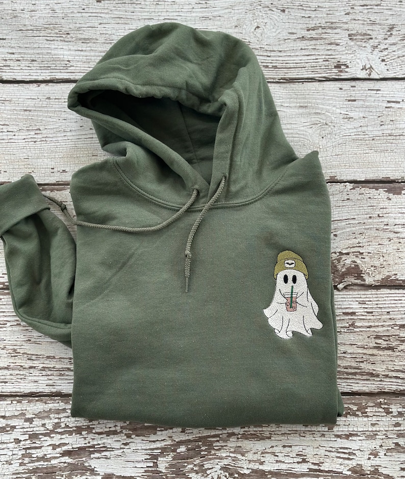 May include: Olive green hoodie with a drawstring hood and a front pocket. The hoodie features an embroidered patch of a ghost wearing a beanie and holding a drink. The ghost is white, the beanie is tan, and the drink is in a clear cup.