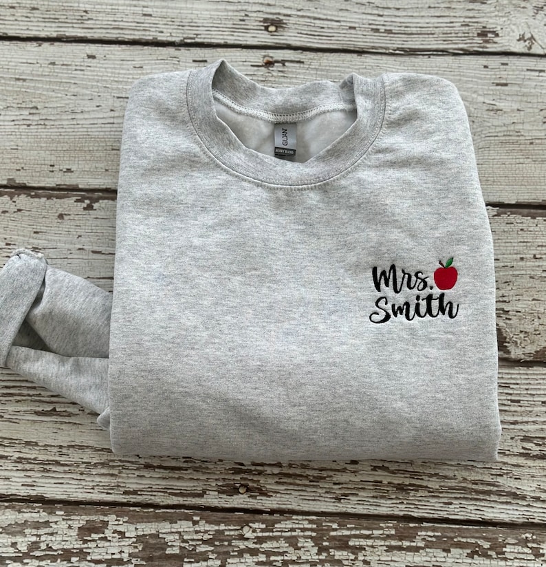 May include: Gray sweatshirt with a black embroidered design that says "Mrs. Smith" with a red apple next to the name.