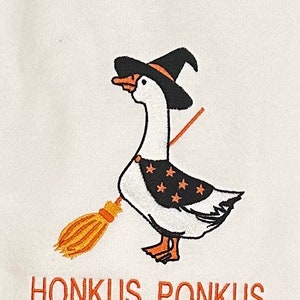 Puede incluir: Un ganso blanco con un sombrero de bruja negro y sosteniendo una escoba. El ganso tiene estrellas naranjas y negras en el pecho. El texto "Honkus Ponkus" est&aacute; escrito debajo del ganso.