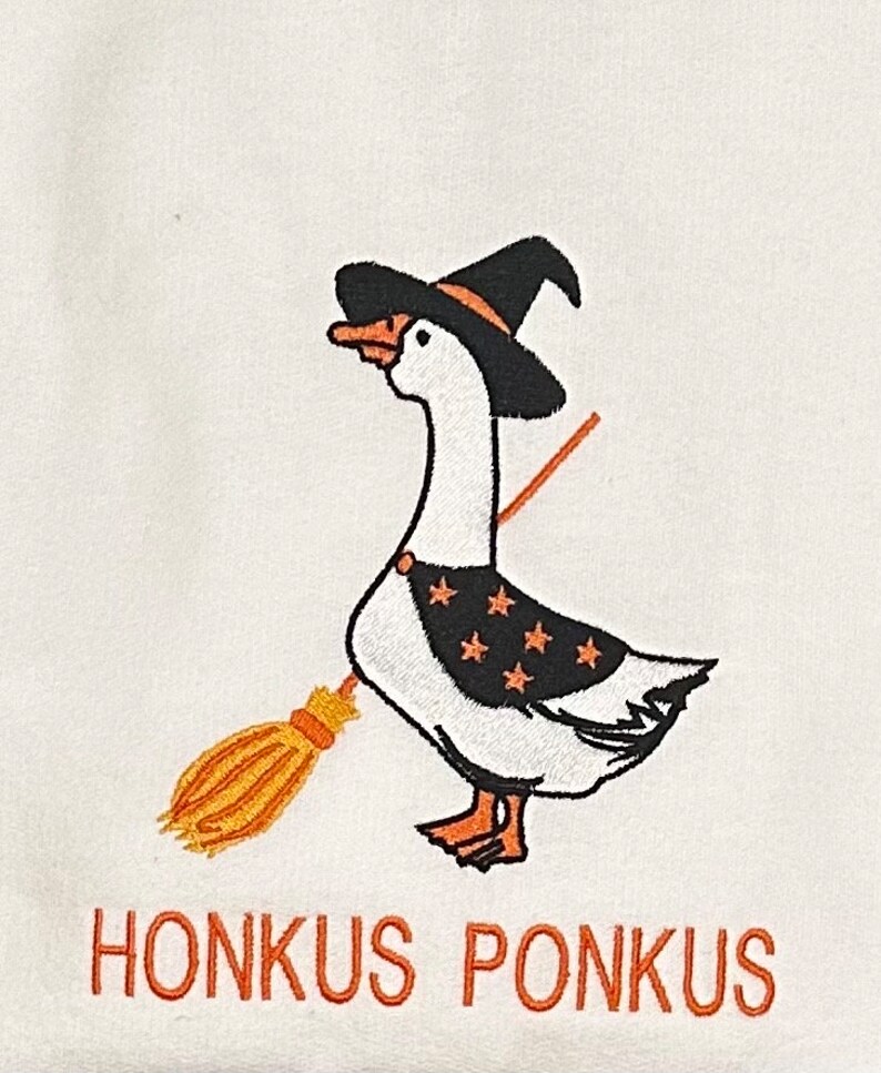 Puede incluir: Un ganso blanco con un sombrero de bruja negro y sosteniendo una escoba. El ganso tiene estrellas naranjas y negras en el pecho. El texto "Honkus Ponkus" est&aacute; escrito debajo del ganso.