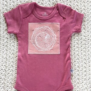 Puede incluir: Un body de bebé rosa polvoriento con mangas cortas. Un parche cuadrado en la parte delantera presenta un diseño geométrico blanco sobre un fondo rosa. El body tiene cierres de presión y la marca "HONEST" es visible.