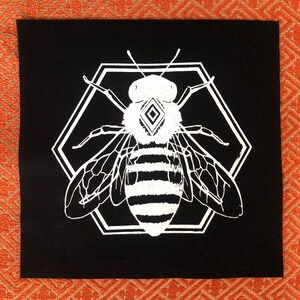 Może przedstawiać: Czarny łata przedstawiająca białą pszczołę z czarnymi paskami. Pszczoła znajduje się wewnątrz białego sześciokąta z diamentowym wzorem w środku głowy pszczoły.