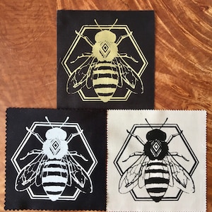 Może przedstawiać: Trzy łatki z tkaniny z motywem pszczoły. Pszczoła znajduje się w sześciokątnym kształcie i ma diament na głowie. Łatki są czarne, białe i beżowe.