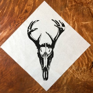 May include: Black and white illustration of a deer skull with large antlers on a white square cloth. The cloth is on a brown wooden surface.