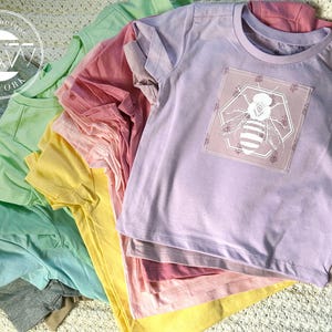 Puede incluir: Una pila de camisetas coloridas en varios tonos pastel, incluyendo lavanda, amarillo y rosa. La camiseta superior es lavanda con un gráfico de abeja. Las camisetas son de material suave y probablemente para niños.