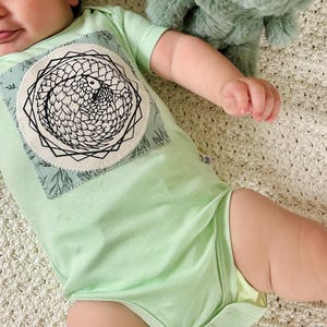 Puede incluir: Un body de bebé verde claro con un parche blanco que presenta una ilustración en blanco y negro de un pangolín. El body tiene un cierre a presión en la parte inferior.
