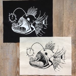 May include: Two fabric patches with a black and white illustration of a deep sea anglerfish. The fish has a large mouth with sharp teeth and a bioluminescent lure.