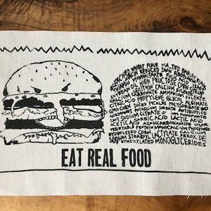 Puede incluir: Una ilustración en blanco y negro de una hamburguesa con el texto "EAT REAL FOOD" debajo. La hamburguesa está compuesta por un pan, una carne, queso, lechuga y tomate. El texto alrededor de la hamburguesa enumera una variedad de ingredientes que se encuentran comúnmente en los alimentos procesados.