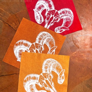 May include: Three fabric squares featuring a white outline of a ram skull with large horns. The squares are red, orange, and brown.