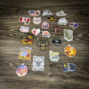 May include: A collection of colorful stickers featuring various designs, including a lineman climbing a pole, a skull with crossed wrenches, and a mermaid with a sunset. The stickers are on a wooden surface.