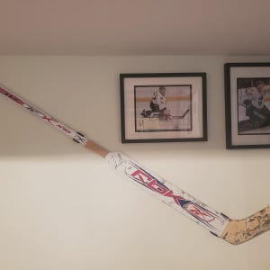 Op de afbeelding: Een witte en rode hockeystick met een zwart en blauw Reebok-logo. De stick is gesigneerd door een hockeyer. De stick is gemonteerd aan een muur met twee ingelijste hockeyfoto's.