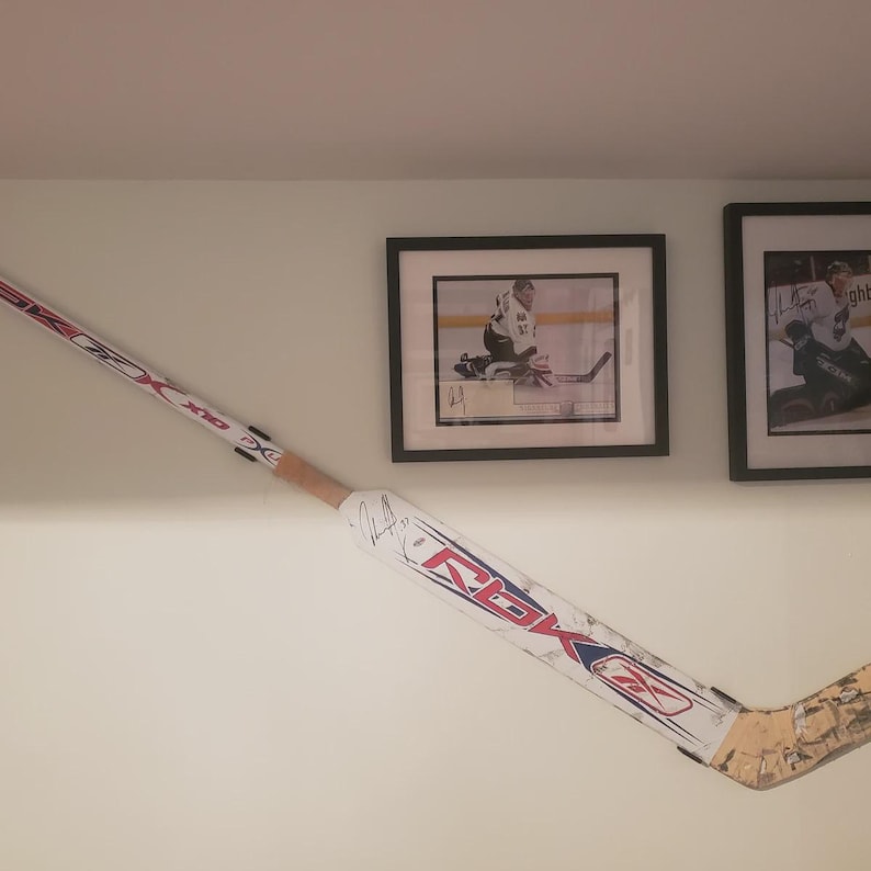 Op de afbeelding: Een witte en rode hockeystick met een zwart en blauw Reebok-logo. De stick is gesigneerd door een hockeyer. De stick is gemonteerd aan een muur met twee ingelijste hockeyfoto's.