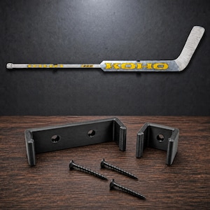 Peut inclure: Une crosse de hockey sur glace KOHO 590 argentée et jaune montée horizontalement au-dessus de deux supports muraux en plastique noir en forme de U et de trois vis métalliques noires. La marque KOHO est inscrite en jaune sur la crosse.