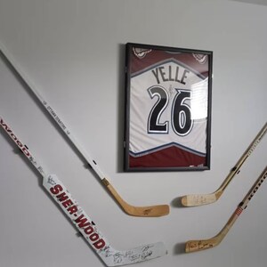 Op de afbeelding: Vier hockeysticks gemonteerd aan een witte muur. Drie van de sticks zijn wit met zwarte accenten en &eacute;&eacute;n is bruin. De sticks zijn gesigneerd met zwarte inkt. Een ingelijst hockeytrui met nummer 26 en de naam "Yelle" hangt aan de muur.