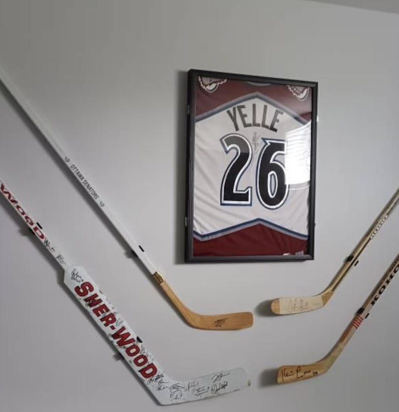 Op de afbeelding: Vier hockeysticks gemonteerd aan een witte muur. Drie van de sticks zijn wit met zwarte accenten en &eacute;&eacute;n is bruin. De sticks zijn gesigneerd met zwarte inkt. Een ingelijst hockeytrui met nummer 26 en de naam "Yelle" hangt aan de muur.