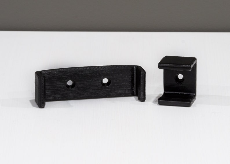 Op de afbeelding: Twee zwarte kunststof wandbeugels met gaten voor schroeven. De beugels zijn ontworpen om een plank of ander object te dragen.