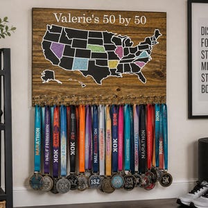 May include: A rectangular wooden wall display featuring a map of the United States with the text "Valerie's 50 by 50" at the top. The map includes black states with some highlighted in color. Below the map are metal hooks holding numerous colorful medals.