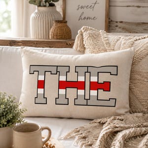 May include: A rectangular beige throw pillow featuring the word THE in large block letters. The letters are outlined in black and filled with a horizontal pattern of grey, white, and red stripes. The fabric has a visible woven texture.