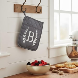 May include: A grey quilted oven glove with a white letter "B" and laurel wreath design hangs on a wooden hook. A white stand mixer, a bowl of berries, and cookies on a wooden board are also visible.