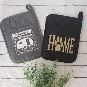 May include: Two gray and black oven mitts with white and gold text. The gray mitt says "Home is wherever we park it" with a white camper van graphic. The black mitt says "HOME" with a gold paw print.