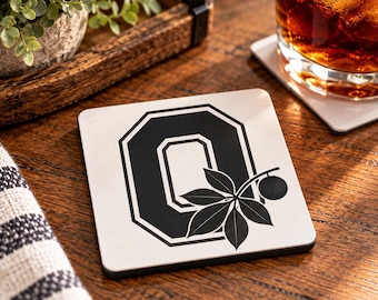 May include: A square white coaster featuring a bold black letter O with a double-line border. A black graphic of a buckeye leaf and a single nut is positioned over the lower right side of the letter O, creating a distinct contrast.