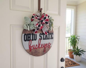 May include: A round wooden door sign featuring the text OHIO STATE in black block letters and buckeyes in red script. The sign has horizontal stripes of dark wood, light gray, and medium wood, topped with a large bow in red, black, and white.