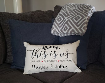 May include: A white throw pillow with black text that reads "This is us... Our life ♥ Our story ♥ Our home Hayley & Lukas" on a gray couch with a gray and white patterned throw blanket.